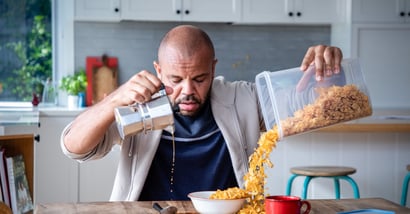 Man pours coffee on the table and cereal on the table while he has his eyes closed looking tired